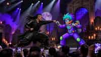 Fighter One VERSUS Fighter Two — a non-stop one-on-one martial-arts FIGHT where the two characters battle each other the entire time using their own unique powers, moves, and fighting style. Fast, precise choreography: punches, blocks, spinning kicks, dodges, throws, and special moves connecting at impressive speed, dust kicking up from the stage floor. EVERY scene is pure mid-combat action — NO preparation, posing, walking up, talking, or spoken dialog. Performed on a raised theater stage with cemetery built as an elaborate scenic backdrop/set, like a Las Vegas stage show with dramatic stage lighting and spotlights. POV HANDHELD PHONE-CAMERA shot filmed by someone in the audience, raised above other people's heads in the crowd, frame slightly unsteady, shot from a fixed audience position. A large crowd fills the FOREGROUND — dark-haired heads silhouetted, many holding up phones recording the show — with shallow focus on the elevated stage where the fight happens, above and in front of the crowd. One continuous handheld audience shot, no cuts to another location; the crowd gasps and cheers. PHOTO STYLE: looks like a real amateur smartphone photo taken from the crowd — candid and unposed, natural daylight, slight handheld motion blur and sensor noise, shallow focus on the stage, photorealistic documentary snapshot, NOT a glossy cinematic studio render. Storyboard cover art