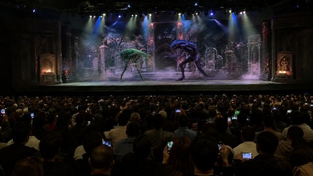Fighter One VERSUS Fighter Two — a non-stop one-on-one martial-arts FIGHT where the two characters battle each other the entire time using their own unique powers, moves, and fighting style. Fast, precise choreography: punches, blocks, spinning kicks, dodges, throws, and special moves connecting at impressive speed, dust kicking up from the stage floor. EVERY scene is pure mid-combat action — NO preparation, posing, or walking up. The fighters MAY exchange short taunts or battle cries in English while fighting, but they never pause or slow down to talk — the combat stays continuous through any dialog. Performed on a raised theater stage with cemetery built as an elaborate scenic backdrop/set, like a Las Vegas stage show with dramatic stage lighting and spotlights. WIDE ESTABLISHING SHOT, camera far back at the rear of the audience and ZOOMED OUT so the ENTIRE stage and a large portion of the crowd are visible in a wide widescreen frame — the fighters appear small in the distance on the full stage, NOT a close-up. POV handheld phone-camera filmed from the audience, raised above other people's heads, frame slightly unsteady, fixed position. A large crowd fills the whole FOREGROUND — many dark-haired heads silhouetted, holding up phones recording the show — with the full elevated stage and the fight clearly in the distance above and in front of them. One continuous handheld audience shot, no cuts to another location; the crowd gasps and cheers. PHOTO STYLE: looks like a real amateur smartphone photo taken from the crowd — candid and unposed, natural daylight, slight handheld motion blur and sensor noise, shallow focus on the stage, photorealistic documentary snapshot, NOT a glossy cinematic studio render. Storyboard